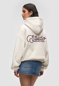 Kremowa bluza z dużym burgundowym haftem "Studio" z tyłu, o luźnym kroju i kapturem ze sznurkiem, zestawiona z dżinsową spódnicą.