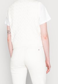 Vitt ribbat stickat väst med flätat mönster, bärs över en vit t-shirt med kort ärm. Par av vita figurnära jeans med bakfickor synliga.