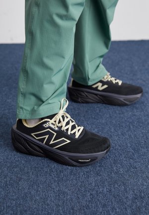 Jambes portant un pantalon vert et des baskets New Balance Fresh Foam noires avec des lacets beige, debout sur un tapis bleu.