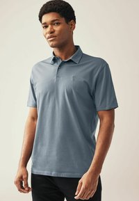 Polo en azul pizarra, hecho de algodón, con un cuello clásico, mangas cortas y un pequeño emblema en el lado izquierdo del pecho.