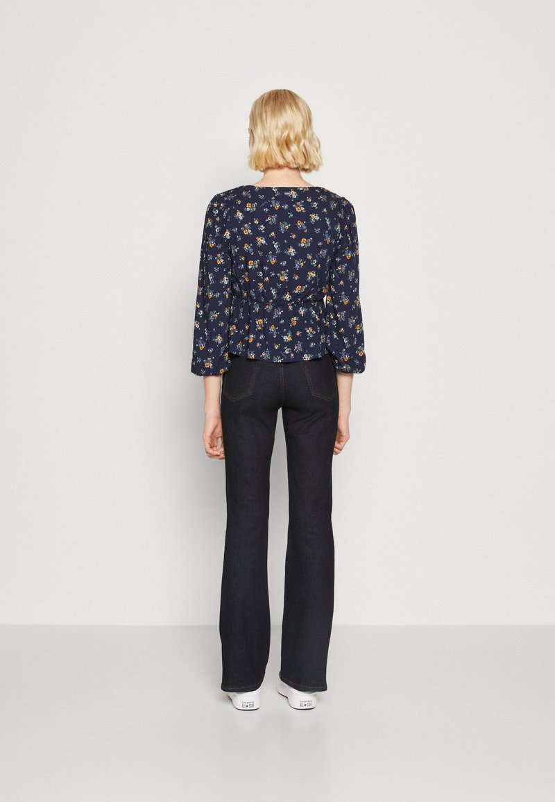 Blusa floreale blu marina con maniche lunghe e vita a peplo, abbinata a jeans svasati scuri e sneakers bianche, vista da dietro.