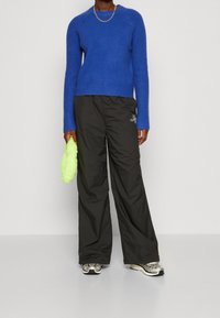 Pull en tricot bleu, pantalon noir aux jambes larges, et baskets argentées. Tient un accessoire duveteux vert clair. Design et textures simples présents.