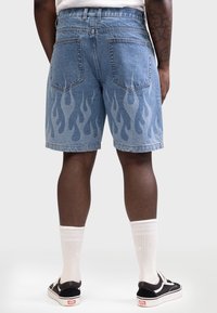 Uomo con pantaloncini di denim blu con motivo a fiamma sul retro, calzini bianchi e scarpe da ginnastica nere, in piedi su sfondo bianco.