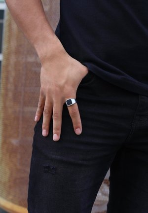 Silver ring with a square black stone, worn on a slender hand. Dark jeans with subtle distressing are visible in the frame.