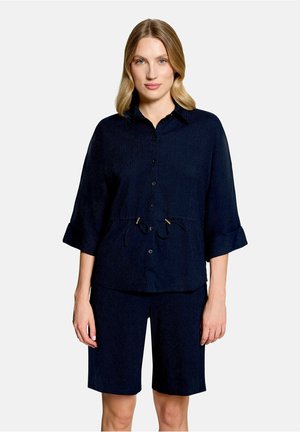 Femme aux cheveux blonds mi-longs, portant une chemise noire boutonnée avec une taille à cordon et un short assorti longueur genou.