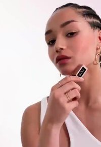 La modelo sostiene un producto de belleza rectangular con una etiqueta negra. Lleva una camiseta blanca y múltiples anillos y aretes de oro. Piel suave, cabello trenzado.