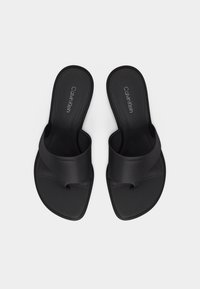 Ζευγάρι μαύρες σαγιονάρες Calvin Klein με φαρδιές λουρίδες και θηλιές για τα δάχτυλα, με επίπεδες σόλες και τετράγωνες μύτες, απεικονίζονται από πάνω σε λευκό φόντο.