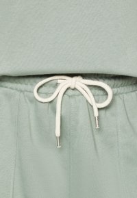 Pantalon de jogging vert clair avec taille à cordon noué en boucle, cordon blanc et embouts en métal argenté, texture douce et ceinture élastique.