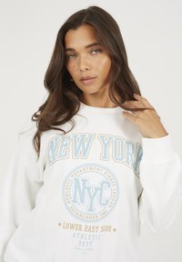 Donna con lunghi capelli castani che si tocca i capelli, indossando una felpa bianca con testo blu e beige "New York NYC Lower East Side Athletic Dept".