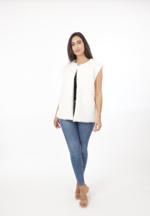 Vrouw draagt een wit mouwloos bontvest, zwart topje, blauwe skinny jeans en doorzichtige hoge hakken, staand tegen een witte achtergrond.