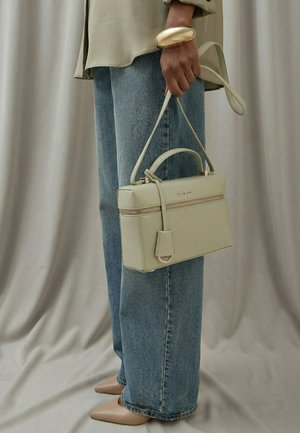 Person trägt beigefarbene Bluse, blaue Jeans, beigefarbene spitze Schuhe, Goldarmband und hält eine hellgrüne Handtasche mit Reißverschluss und Griffen oben.