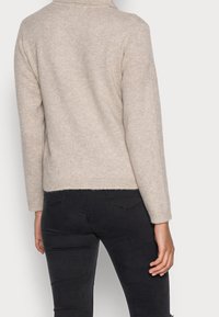 Beige gebreide trui met een hoge kraag, geribbelde manchetten en onderrand. Korte pasvorm, zachte textuur, gedragen met zwarte strakke broeken.