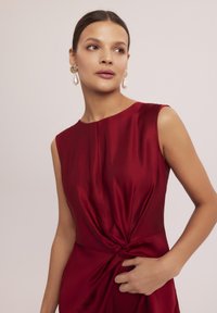 Czerwona satynowa sukienka bez rękawów z węzłowym detalem w talii. Gładka faktura i eleganckie drapowanie podkreślają rzeźbioną sylwetkę.