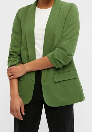 Personne portant un blazer vert avec les manches retroussées sur une chemise blanche et un pantalon noir, les bras croisés à la taille.