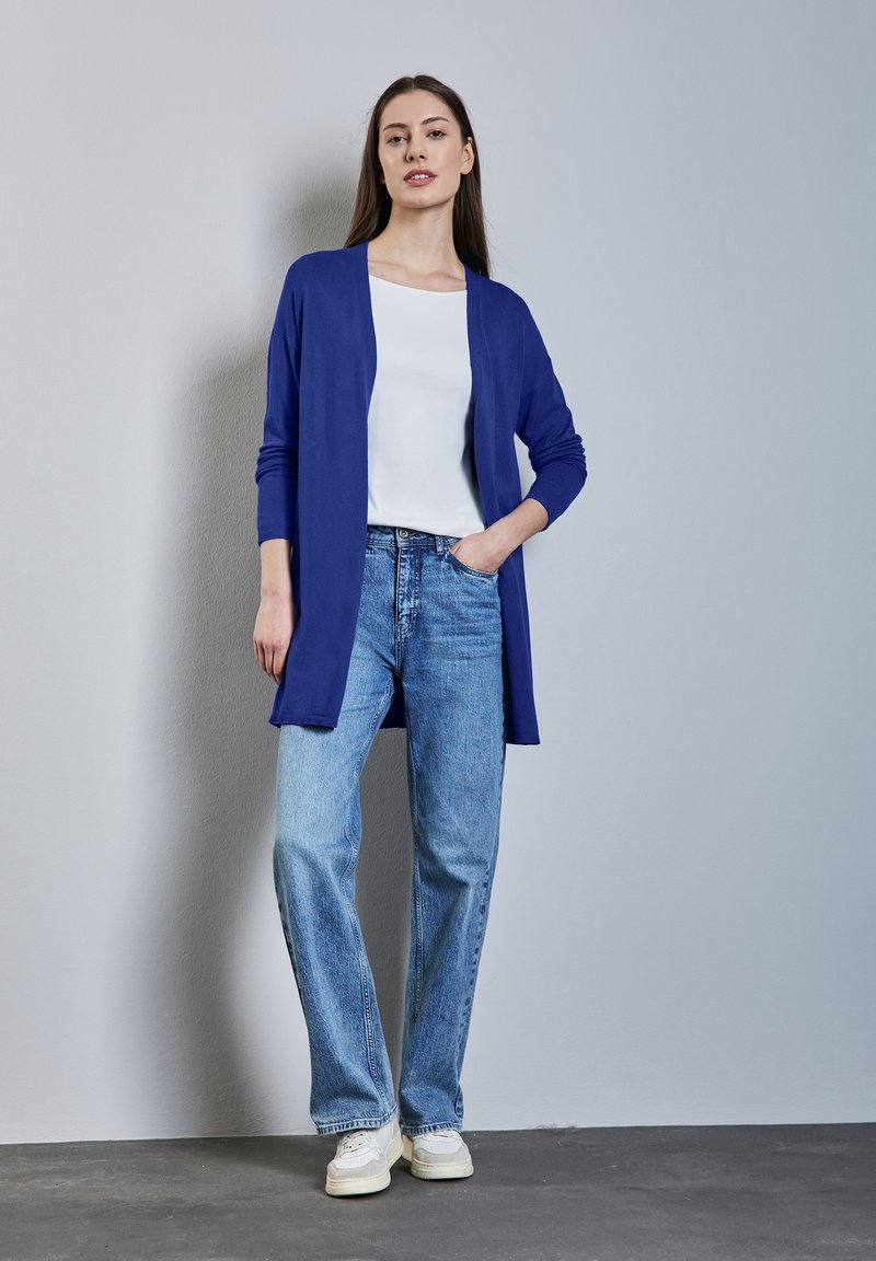 Långärmad blå cardigan över en vit skjorta, i kombination med ljusblå vida jeans och vita sneakers, mot en neutral bakgrund.