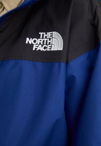 Plava i crna jakna glatke teksture, s istaknutim bijelim logom "THE NORTH FACE" na području lijevog ramena.