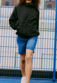 Svart huvtröja med en framficka, tillsammans med blå denimshorts. Bärs med vita sneakers, stående mot ett blått staket.