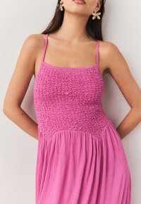 Lyserød kjole med smocked krop, tynde stropper og flydende nederdel. Har tekstureret stof og rynkede designdetaljer.