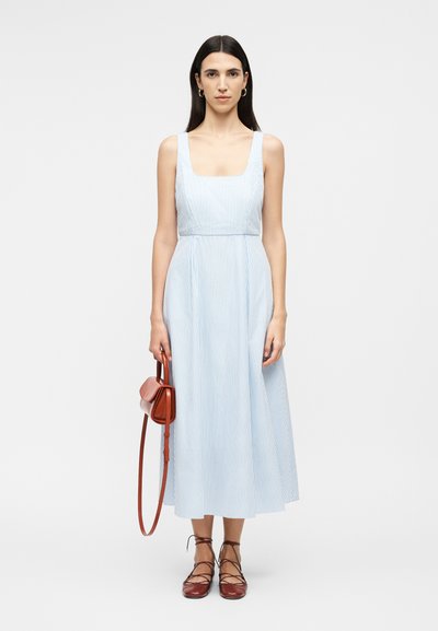 Žena oblečená v svetlomodrom a bielom pruhovanom midi šatách bez rukávov, držiaca hnedú kabelku cez rameno a obutá v hnedých teniskách na šnurovanie.