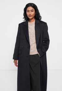 Cappotto lungo di lana scura con ampi risvolti e chiusura doppiopetto, indossato sopra un maglione chiaro a trecce color crema e pantaloni scuri.