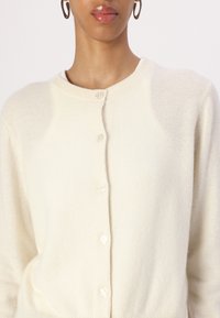 Cardigan couleur crème en tissu doux, avec un col rond, une fermeture à boutons sur le devant et des poignets côtelés.