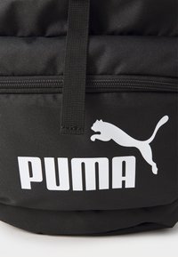 Juodas audinio kuprinė, išsiskirianti ryškiu baltu Puma logotipu ir ženklinimu, su lygiu paviršiumi ir viršutiniu kilpu nešimui. Matomas užtrauktuko detalizavimas.