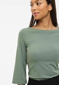 Frau mit langen dunklen Haaren, die kleine Perlenohrringe, ein hellgrünes, schiffhalsförmiges Oberteil mit dreiviertel Ärmeln und schwarze Hosen trägt, blickt nach rechts.