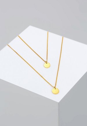 Deux délicats colliers chaîne en or avec des pendentifs ronds en or présentés sur une surface blanche inclinée sur un fond gris.