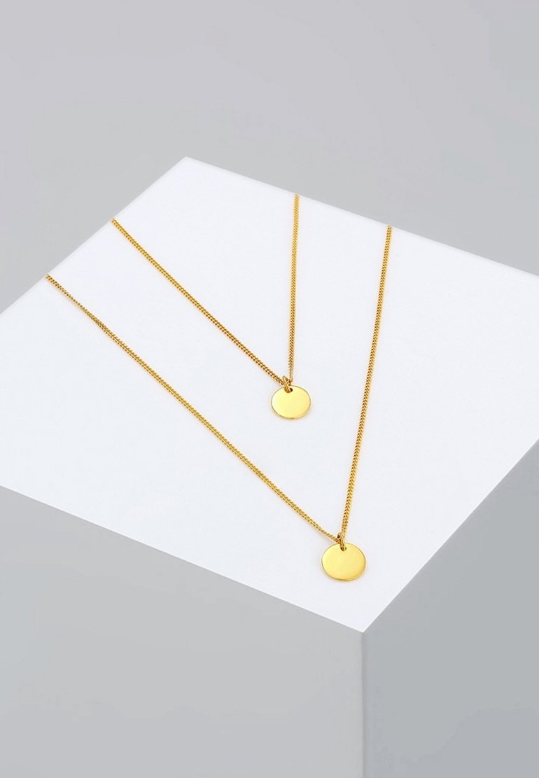 Deux délicats colliers chaîne en or avec des pendentifs ronds en or présentés sur une surface blanche inclinée sur un fond gris.