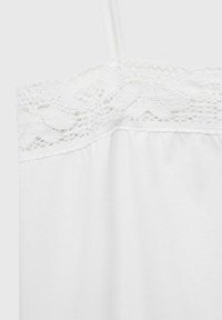 Witte katoenen hemd met een delicate kanten rand langs de bovenrand en dunne schouderbandjes tegen een effen achtergrond.