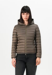 Vrouw met steil donker haar, gekleed in een bruine gewatteerde hooded jack, dichtgeritst, gecombineerd met een zwarte spijkerbroek, staande tegen een witte achtergrond.
