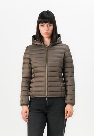 Kvinne med rett, mørkt hår iført en brun vattert hettejakke med glidelås, kombinert med svarte jeans, som står mot en hvit bakgrunn.