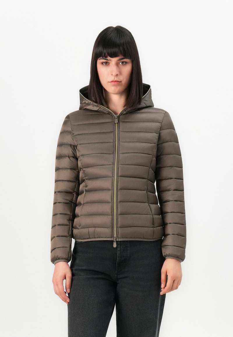 Vrouw met steil donker haar, gekleed in een bruine gewatteerde hooded jack, dichtgeritst, gecombineerd met een zwarte spijkerbroek, staande tegen een witte achtergrond.