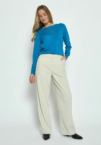 Maglione blu a maniche lunghe abbinato a pantaloni larghi color crema e scarpe nere a punta, che mettono in risalto una texture liscia e una vestibilità rilassata.