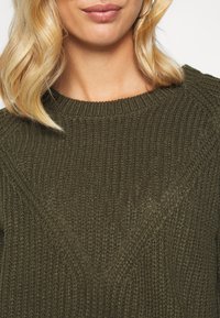 Olivgrüner, gestrickter Pullover mit einem strukturierten Muster, ausgestattet mit einem Rundhalsausschnitt und subtilen diagonalen Rippungen auf der Vorderseite.