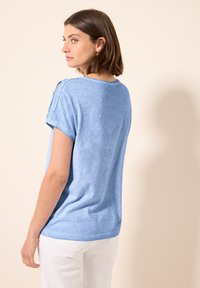 T-shirt azzurro chiaro a maniche corte, morbido al tatto, con scollatura rotonda e maniche arrotolate, dal fit rilassato e leggero lucido, indossato con pantaloni bianchi.