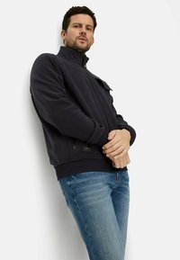 Dunkelblaues Fleece-Jacke mit hohem Kragen, Reißverschluss und Seitentaschen, kombiniert mit hellblauen Jeans. Minimalistisches Design und glatte Textur.