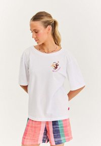 Žena s blond culíkem, oblečená v bílém tričku s potiskem a proužkovaných šortkách v několika barvách, stojí s rukama za zády a dívá se dole vlevo.