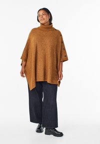 Brązowy dziany ponczo z wysokim kołnierzem, z guzikami po bokach. Połączone z ciemnymi, szerokimi spodniami dżinsowymi i błyszczącymi czarnymi botkami za kostkę.