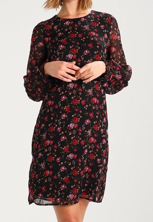 Woman wearing a black knee-length dress with long sleeves and red, purple, and brown floral pattern, hands gently clasped at waist.