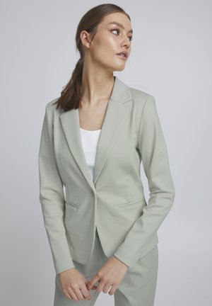 Vrouw met bruin haar in een paardenstaart, die een lichtgrijze blazer draagt over een witte top, kijkt naar rechts tegen een effen grijze achtergrond.