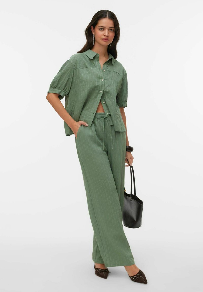 Femme portant une chemise ample verte à fines rayures et un pantalon, tenant un sac à main noir, debout avec une main dans la poche sur fond blanc.