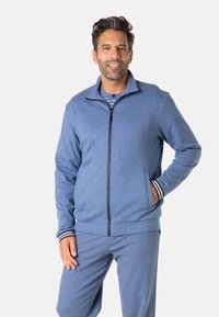 Veste zippée bleue pour homme en matière douce avec poches latérales, poignets côtelés aux rayures blanches et bleu marine, associée à un pantalon assorti.