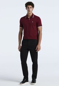Bordeauxfarbener Poloshirt mit weißen Akzentstreifen am Kragen und an den Ärmeln, kombiniert mit schwarzen Slim-Fit-Hosen und schwarzen Sneakers.