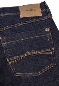 Jeans in denim blu scuro con un motivo di cucitura distintivo sulla tasca posteriore e un'etichetta in pelle beige con la scritta "MUSTANG" in rilievo.