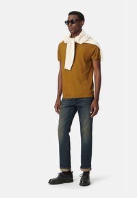 Maglietta marrone a maniche corte indossata con un maglione crema appoggiato sulle spalle, abbinata a jeans in denim scuro e scarpe nere.