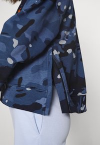 Kamouflagemönstrad jacka i nyanser av blått med grå och svarta detaljer. Har en sidodragkedja och ribbad kant för en avslappnad passform.