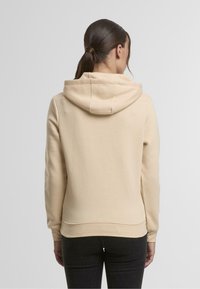 Sudadera con capucha beige de textura suave y bolsillo tipo canguro. Mangas largas y puños de canalé, con detalles de costura en la capucha.
