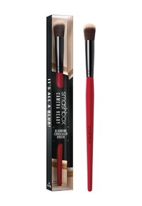 Pędzel do korektora Smashbox Blurring ma stożkowy kształt główki z miękkim włosiem, czerwone rączkę i czarną ferrulę, zapakowany w przezroczysty pudełko.