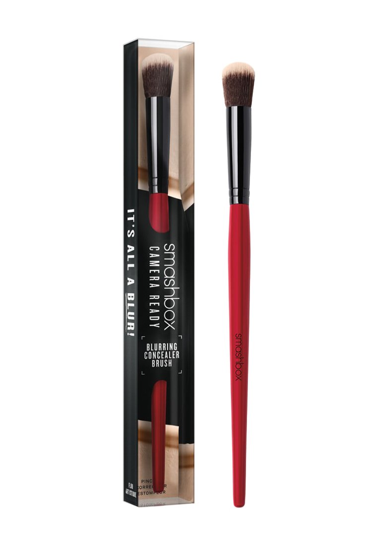 Pędzel do korektora Smashbox Blurring ma stożkowy kształt główki z miękkim włosiem, czerwone rączkę i czarną ferrulę, zapakowany w przezroczysty pudełko.